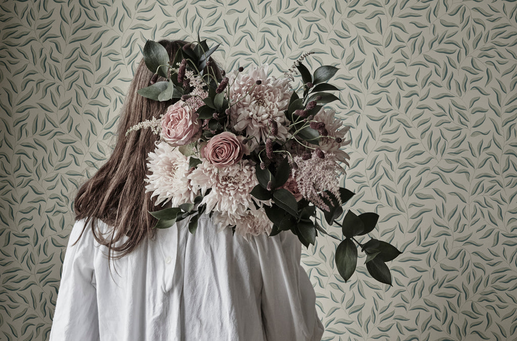 Karolina Foliage, Pattern Wallpaper iin sage featured on a wall of a room with a girl carrying a bouquet of flowers.