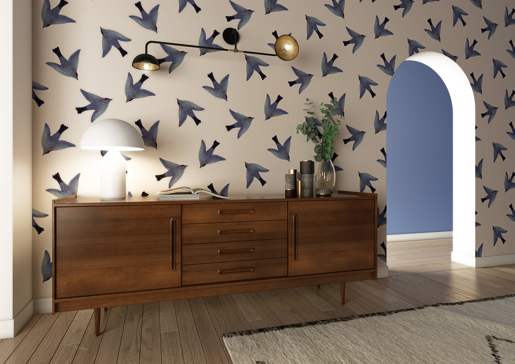 L’oiseau Swallows wallpaper adorns a room with blue bird silhouettes. A mid-century sideboard sits against it, topped with a lamp and decor. A modern light fixture hangs above. The floor is light wood, partially covered by a rug.
