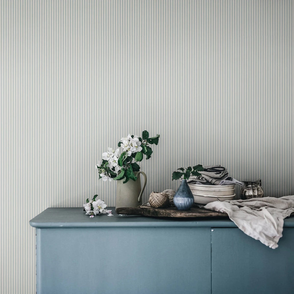 Laura Stripes, Pattern Wallpaper in blue featured on a wall of a room with a cabinet with vase and and cloth on top