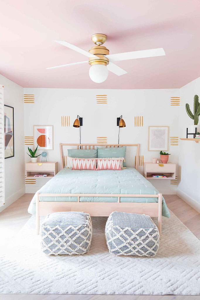 Spacious bedroom with pastel pink walls, adorned with Line Decals, Wall Decals. Features a double bed with a mint green bedspread, gold-accented wall sconces, a white ceiling fan, and woven ottomans.