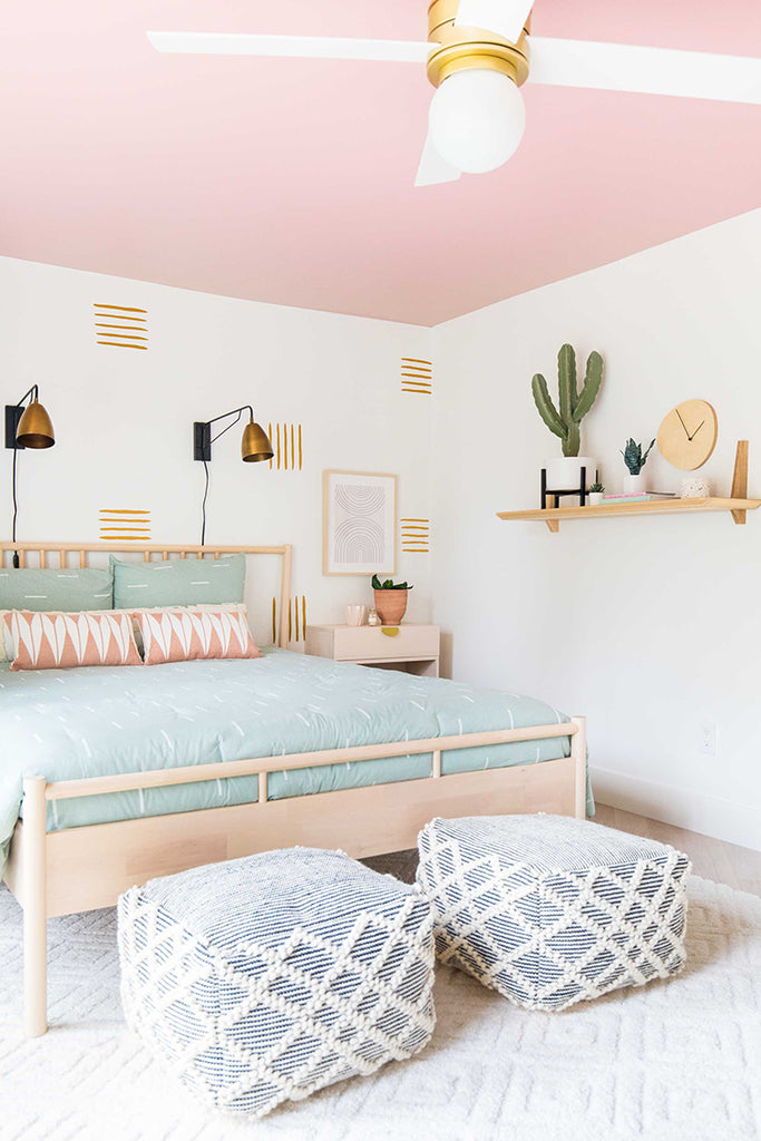 Spacious bedroom with pastel pink walls, adorned with Line Decals, Wall Decals. Features a double bed with a mint green bedspread, gold-accented wall sconces, a white ceiling fan, and woven ottomans.