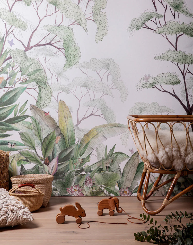 Magical Forest, Mural Wallpaper, adorns a child’s playroom wall. A rattan crib, rattan baskets, and scattered wooden toys complete the scene.