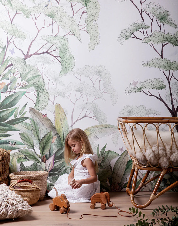 Magical Forest, Mural Wallpaper, graces a child’s playroom wall. A girl is seen playing on the ground, with a rattan crib and baskets nearby. Wooden toys are scattered around the room.