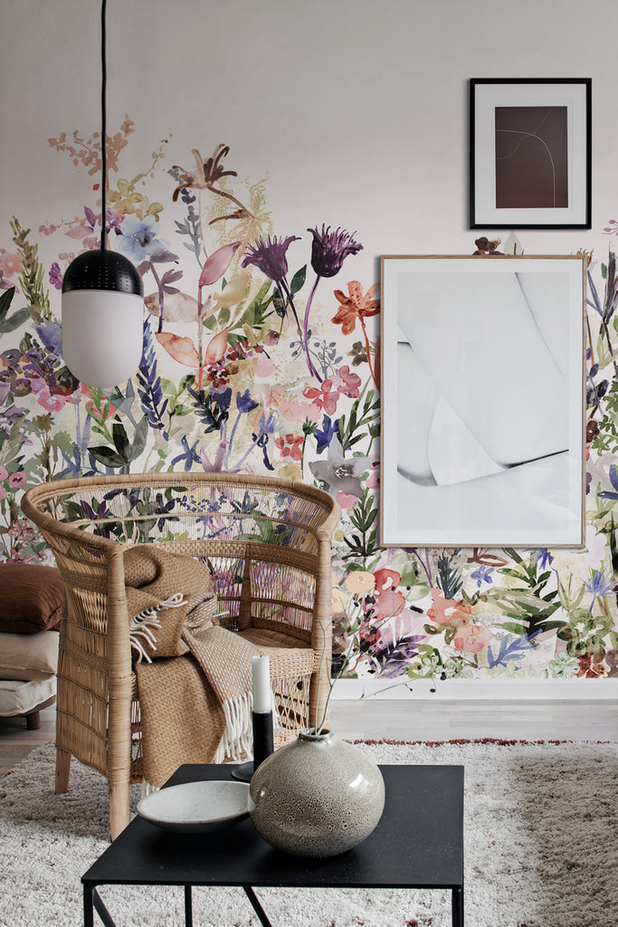 May Meadow, Floral Mural Wallpaper in multicolor pastel featured on a wall of a room with rattan chair and black table with ceramic jar on it