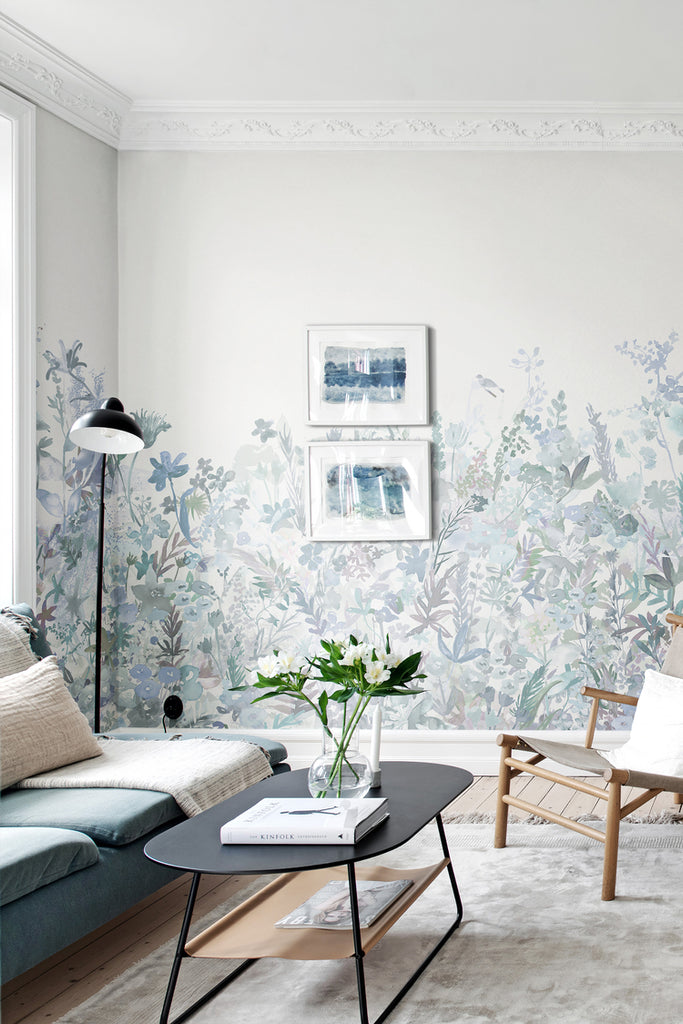 May Meadow, Floral Mural Wallpaper in soft blue featured on a wall of a living room with black table, rattan chair and a dark blue sofa