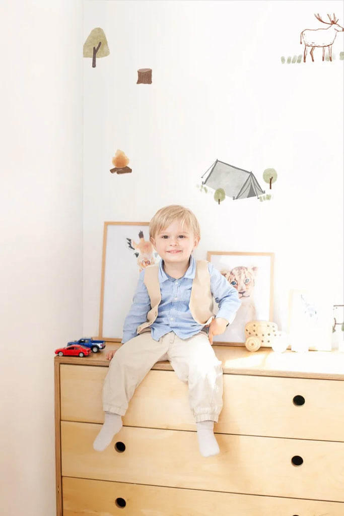 A cheerful boy sits atop a light wooden dresser in a bright, stylish room. The interior features playful decor, including framed animal prints and wooden toys. The mini camper and whimsical Mini Camper, Pattern Wallpaper in White adds a touch of adventure to the clean, modern space.