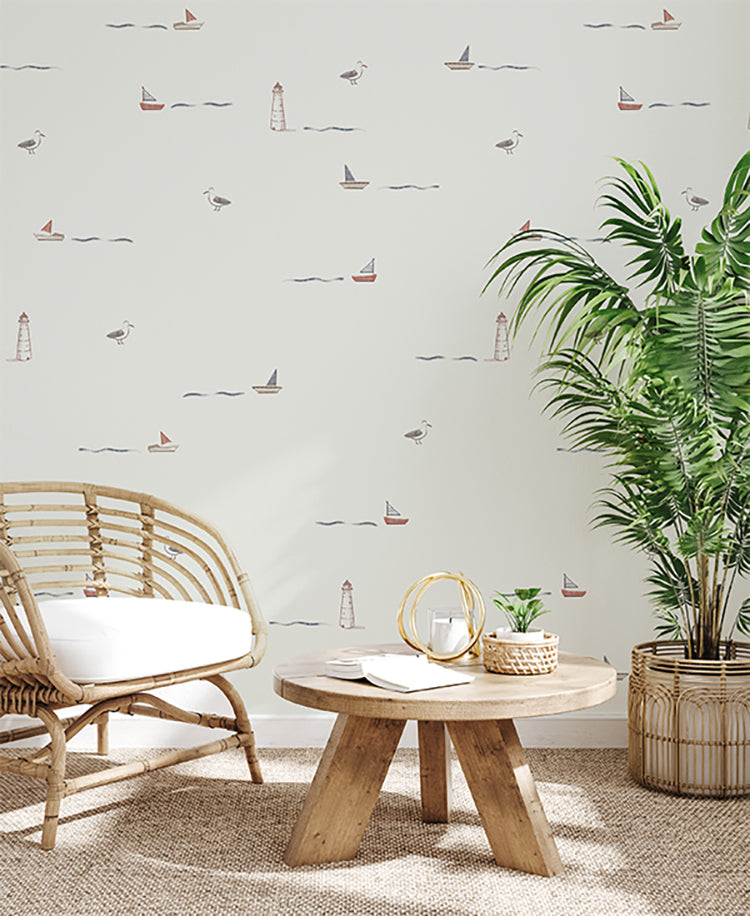 A serene corner of a room, bathed in natural light, showcases a rustic charm with a rattan armchair and wooden table, complemented by the Mini Seafarer, Pattern Wallpaper in Light Grey that adds a maritime touch with its sailboat and lighthouse motifs.