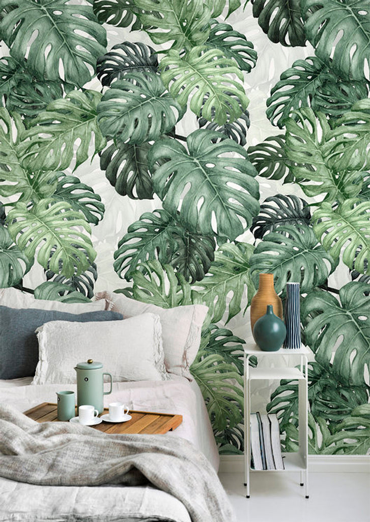 Monstera Green, Tropical Mural Wallpaper featured on a wall of a bedroom and a bed with white sheets and pillows. Next to the bed, there is a side table full of ceramic jars and vases. 