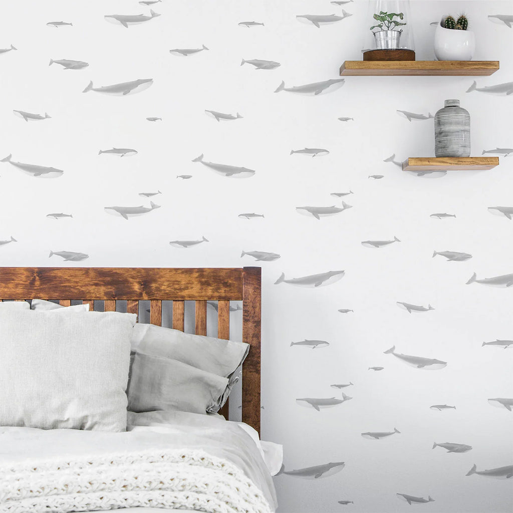 Serene bedroom scene with a wall adorned by ‘Ocean Whale, Pattern Wallpaper’, featuring grey whale silhouettes on a white backdrop. A wooden headboard, cozy bedding, and two wooden shelves with decor items are visible