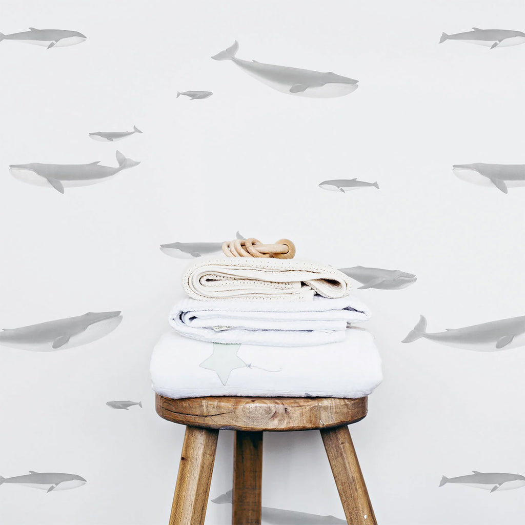 Minimalist ‘Ocean Whale, Pattern Wallpaper’ with grey whale silhouettes on a white backdrop. A wooden stool with neatly folded towels is in the foreground.