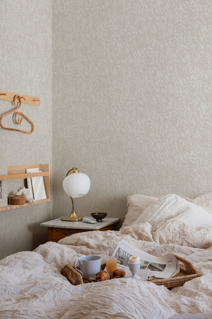 Olof, Floral Pattern Wallpaper in Sand featured on the wall of a bed room, with a bed with soft fabrics and pillows with a side table and an small lamp on it. 