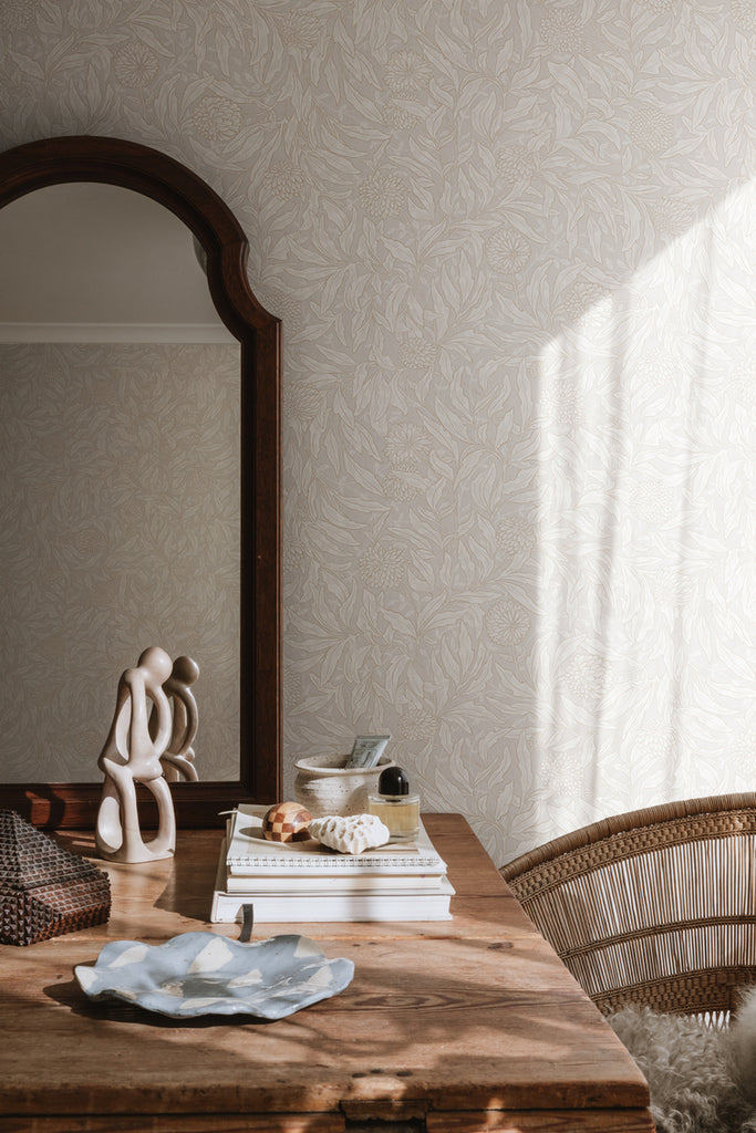 Olof, Floral Pattern Wallpaper in Sand  featured on a wall of a room with a wooden table that ahs books and several items on top of it, beside it is a rattan chair