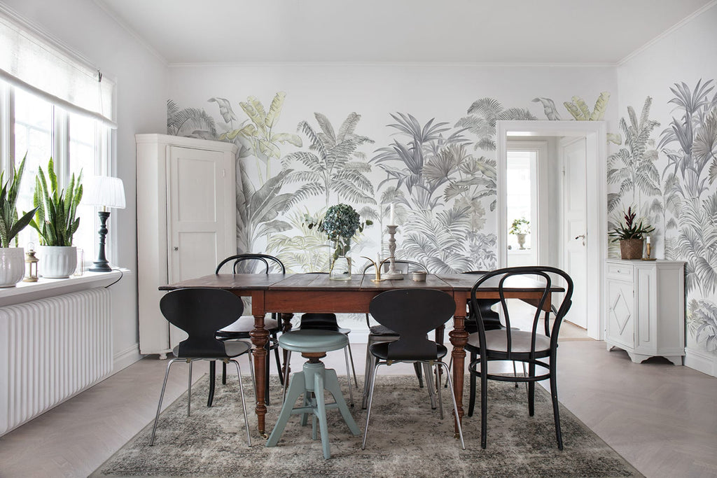 A spacious, well-lit dining room with a large wooden table and chairs, white cabinetry adorned with indoor plants, and a rug on the light floor. The room is enhanced by a Palm Garden, Tropical Mural Wallpaper.