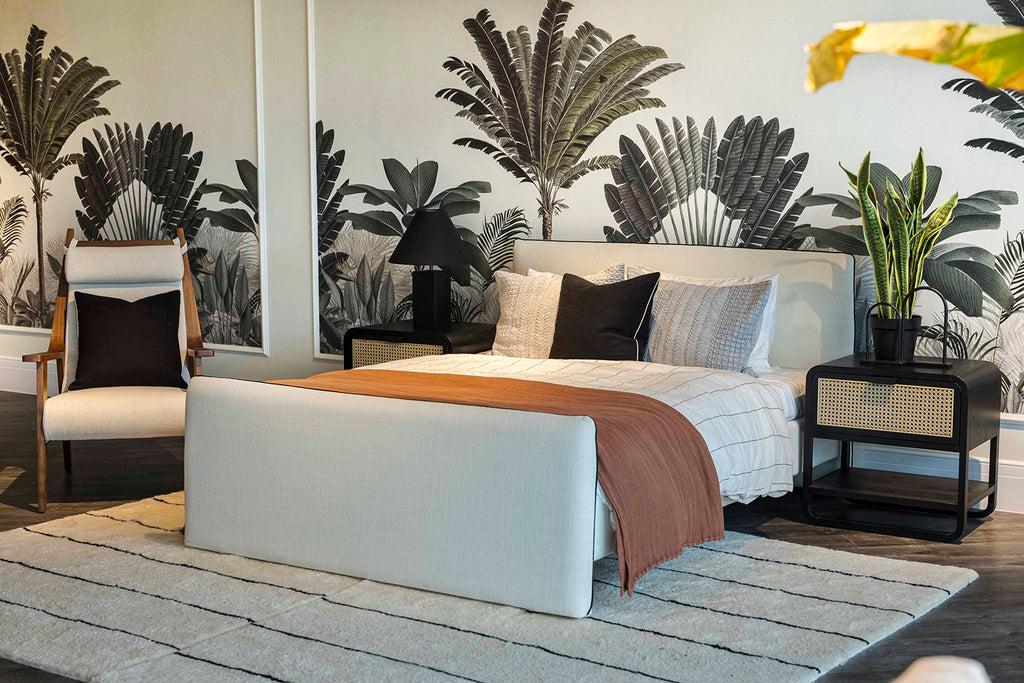 A tranquil bedroom showcasing Palm Paradise, Tropical Mural Wallpaper in Green with large palm leaf patterns. The room features a tidy bed with white and brown linens, complemented by black side tables and contemporary lamps.