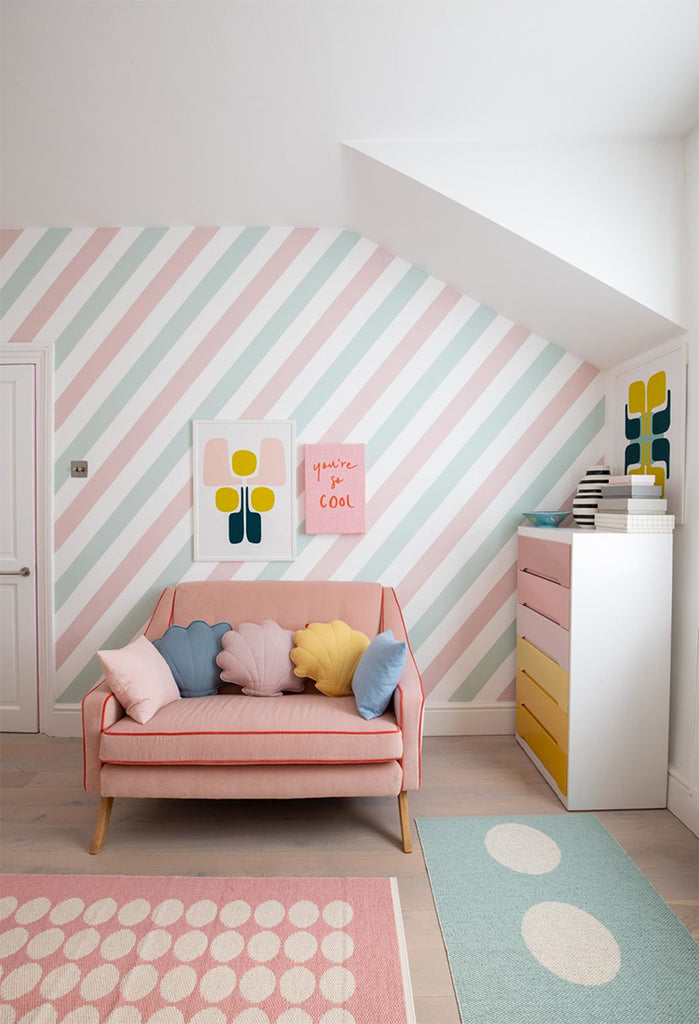 A vibrant attic room with a coral sofa, Pastel Candy Stripes, Wallpaper, colorful throw pillows, contemporary art, a multicolored chest of drawers, and playful area rugs.