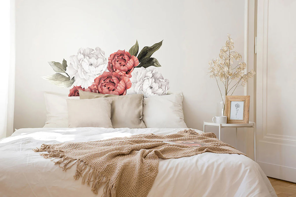 A bedroom with a calming ambiance, featuring a large wall decal of soft pink and white peonies, named Peonies Pack, Floral Wall Decals in Crimson and White, above a neatly made bed with white bedding and a tan throw.