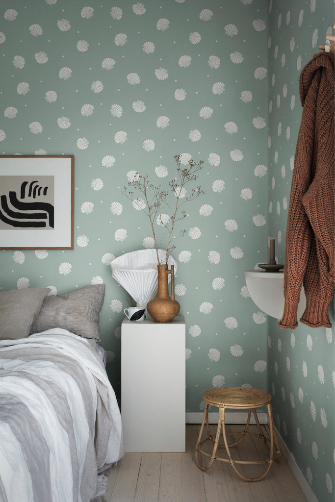 Picking Seashells, Wallpaper in Sage Featured on a wall of a bedroom with a comfortable bed, white side table and a rattan chair