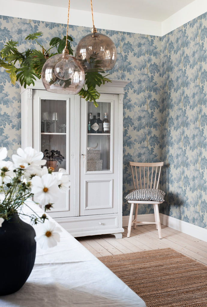 Raphael Floral, Wallpaper in Light Blue featured on a wall of a room with a white kitchenware cabinet and a table with white cloth and a black vase with white flowers