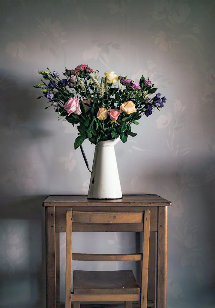 Rebecca, Floral Pattern Wallpaper in Dusky Blue featured on a wall of a room with wooden chair and table, on top of it is a vase with a bouquet of flowers 