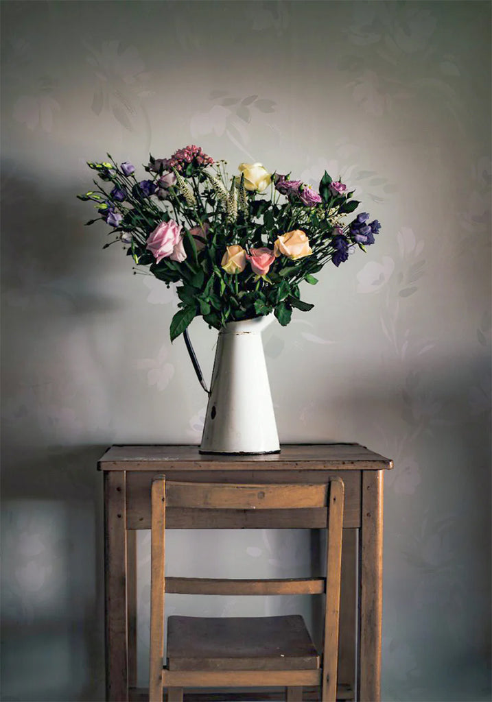 Rebecca, Floral Pattern Wallpaper in Sage featured on a wall of a room with wooden chair and table, on top of it is a vase with a bouquet of flowers 