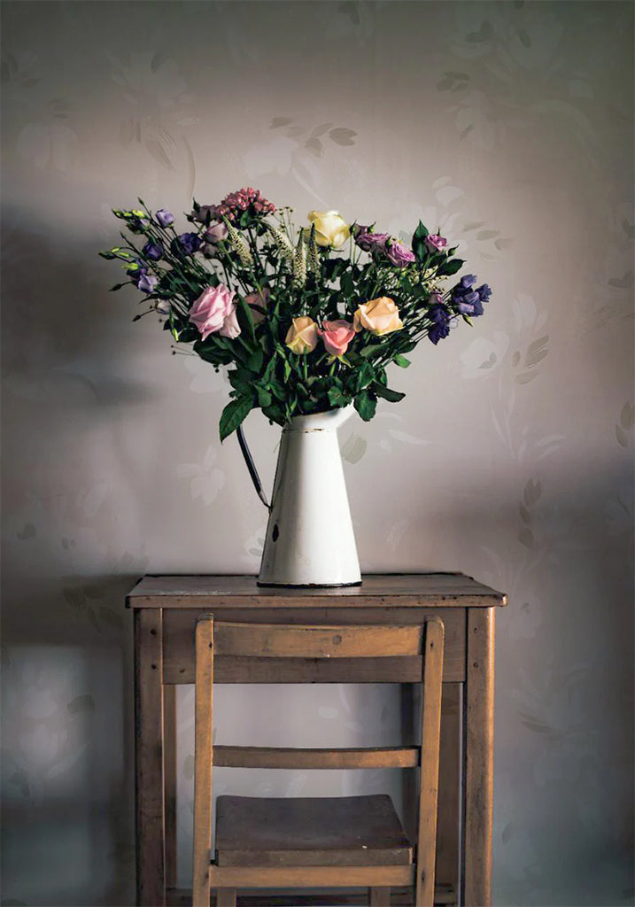 Rebecca, Floral Pattern Wallpaper in Sand featured on a wall of a room with wooden chair and table, on top of it is a vase with a bouquet of flowers 