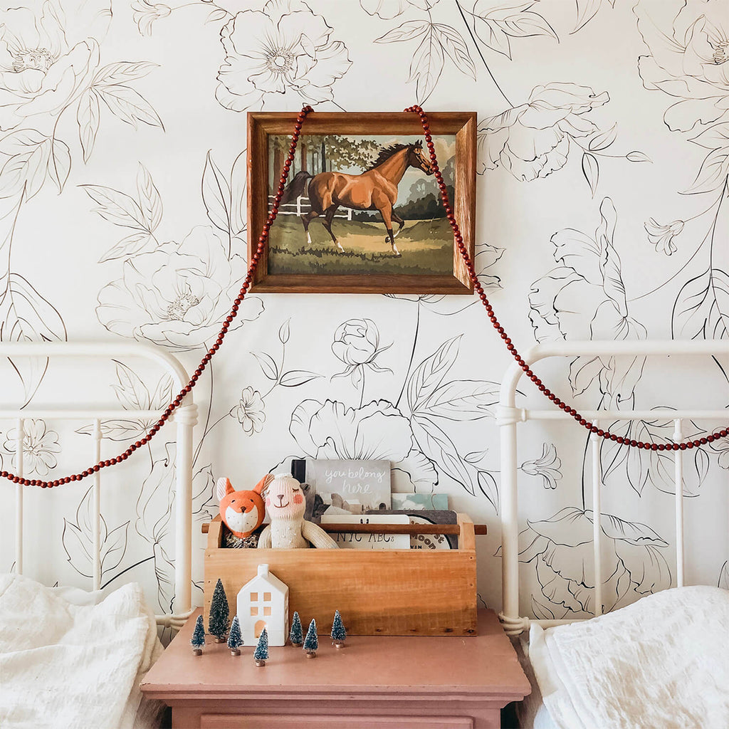 A cozy corner in a room with Renae Delicate Flowers Pattern Wallpaper. A wooden shelf holds books, figurines, and a horse painting. The shelf is placed on a pink surface between two white cushions.