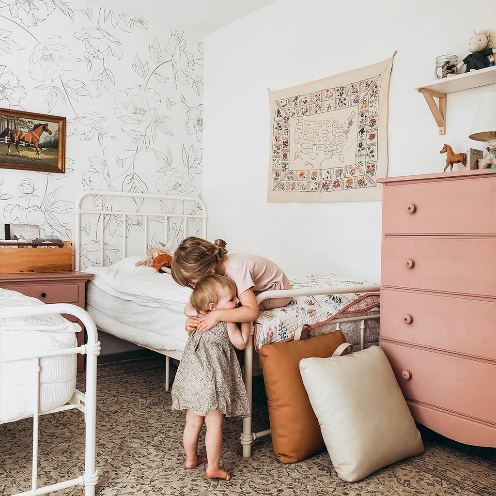 A cozy room with Renae Delicate Flowers wallpaper. A white bed and pink dresser add to the decor. Two people near the bed share a warm embrace, adding a sense of love and comfort to the room.