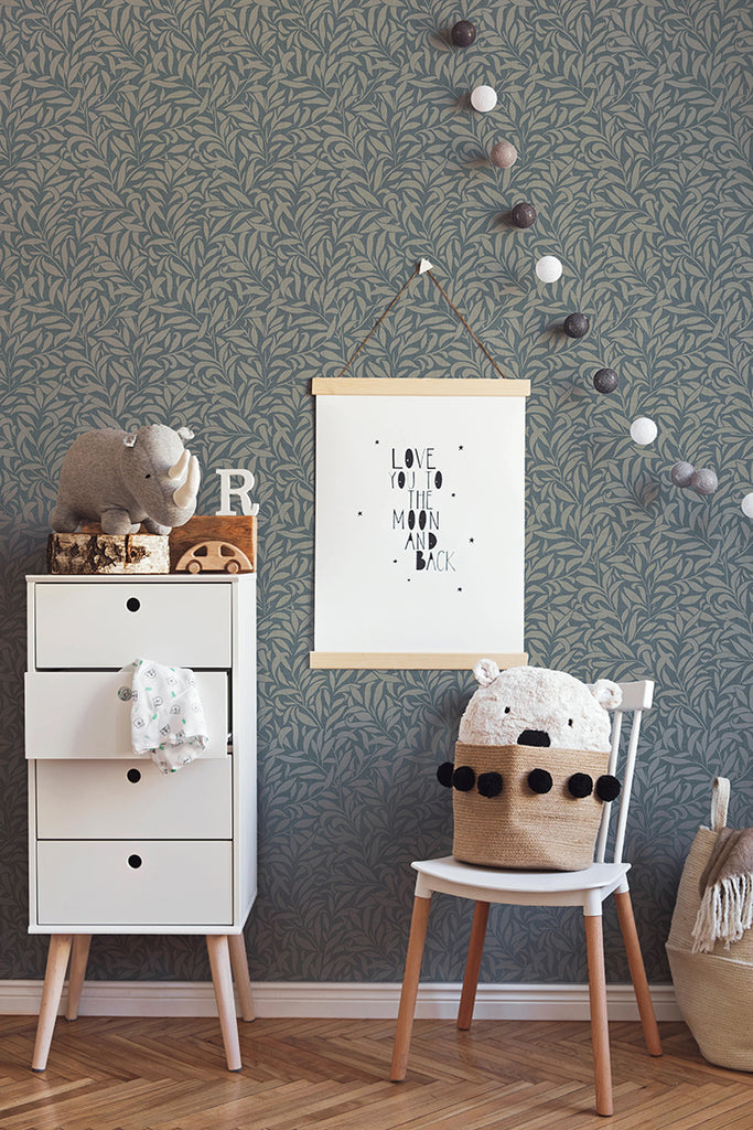 A cozy room bathed in soft light, featuring a white dresser adorned with toys. A chair with a knitted cover invites relaxation, next to a basket filled with comfort items. A framed quote adds a personal touch to the wall. The room is brought to life by the ‘Rippling Leaves’ pattern wallpaper in Blue, adding a touch of nature’s tranquility.