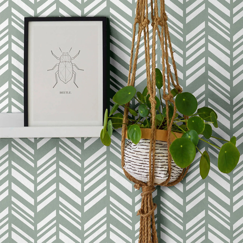 A cozy room with Sage Layered Herringbone Pattern Wallpaper. A framed beetle sketch and a hanging potted plant with lush green leaves add to the room’s decor.
