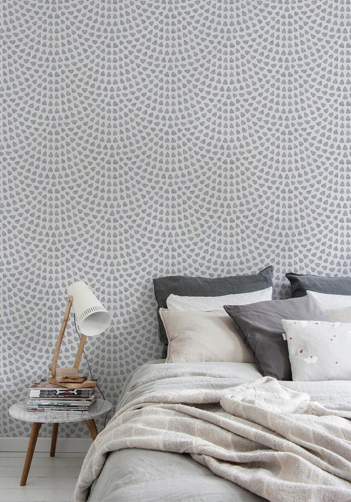 Cozy bedroom with Scallop Polka Dots, Wallpaper. A comfortable bed with gray bedding is in the foreground, next to a wooden bedside table with a lamp and books.