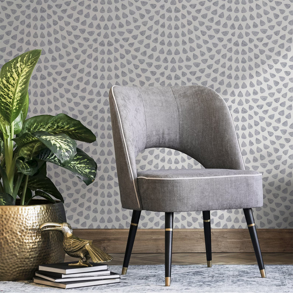 A corner of a room featuring a modern gray upholstered chair with black legs. Beside the chair, a golden pot holds a lush green plant. Underneath the pot, two books are stacked with a small golden bird figurine on top. The room is adorned with Scallop Polka Dots, Wallpaper