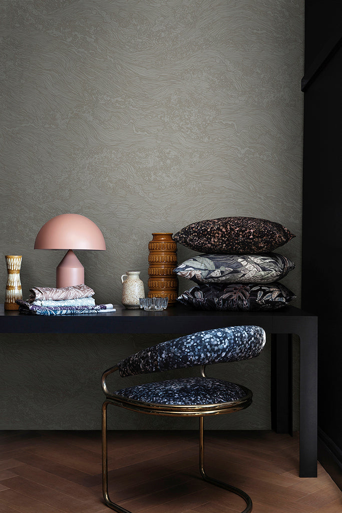 Sea Foam, Wallpaper in Light Grey featured on a wall of a room, with black table that has multiple patterned pillow, and pink lamp, with a patterned chair next to it. 