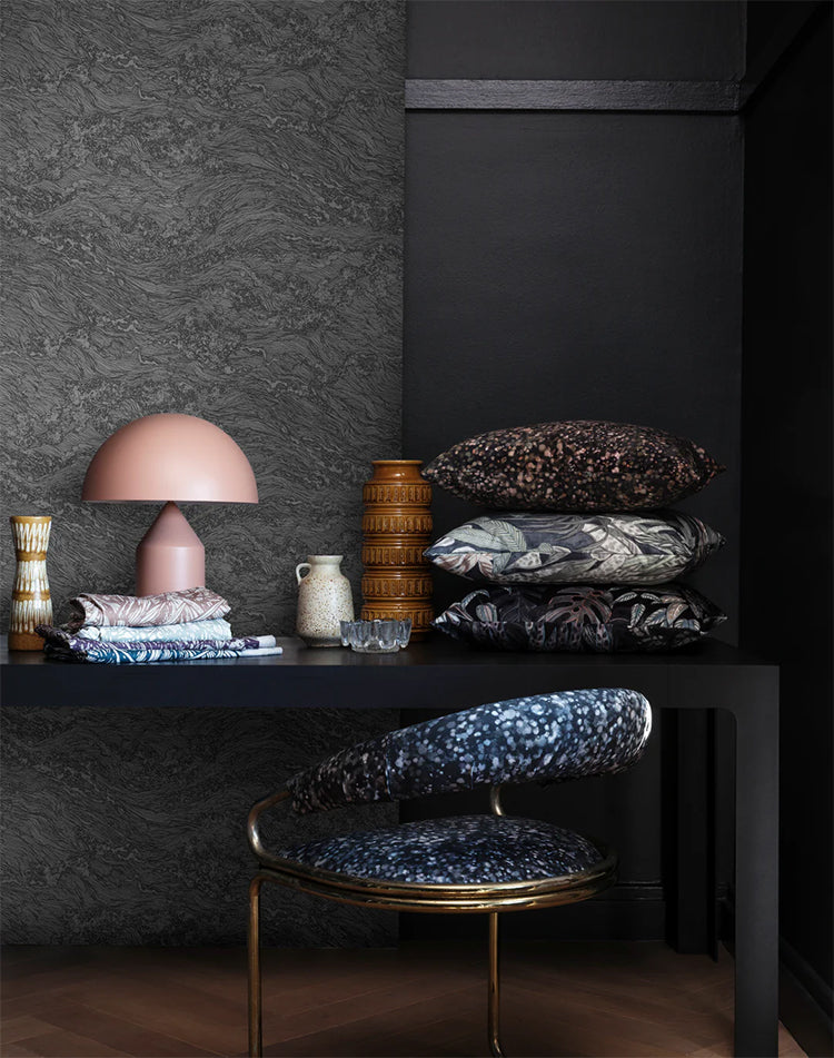 Sea Foam, Wallpaper in Stratos Grey featured on a wall of a room, with black table that has multiple patterned pillow, and pink lamp, with a patterned chair next to it. 
