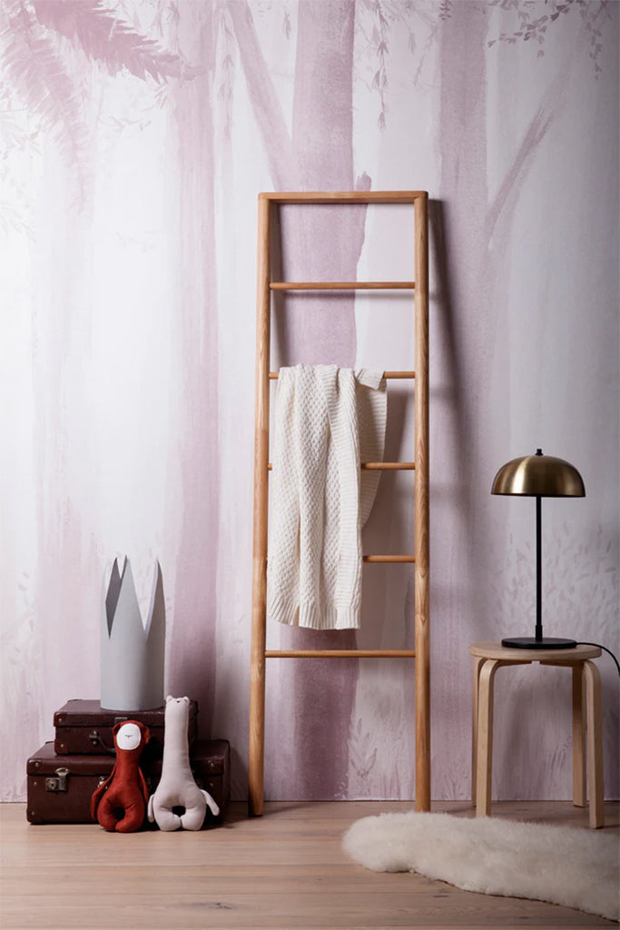 Featuring the Secret Woodland Pink Mural Wallpaper, a room is adorned with a wooden ladder, a towel, a lamp atop a wooden stool, and two stacked brown briefcases beside the ladder.