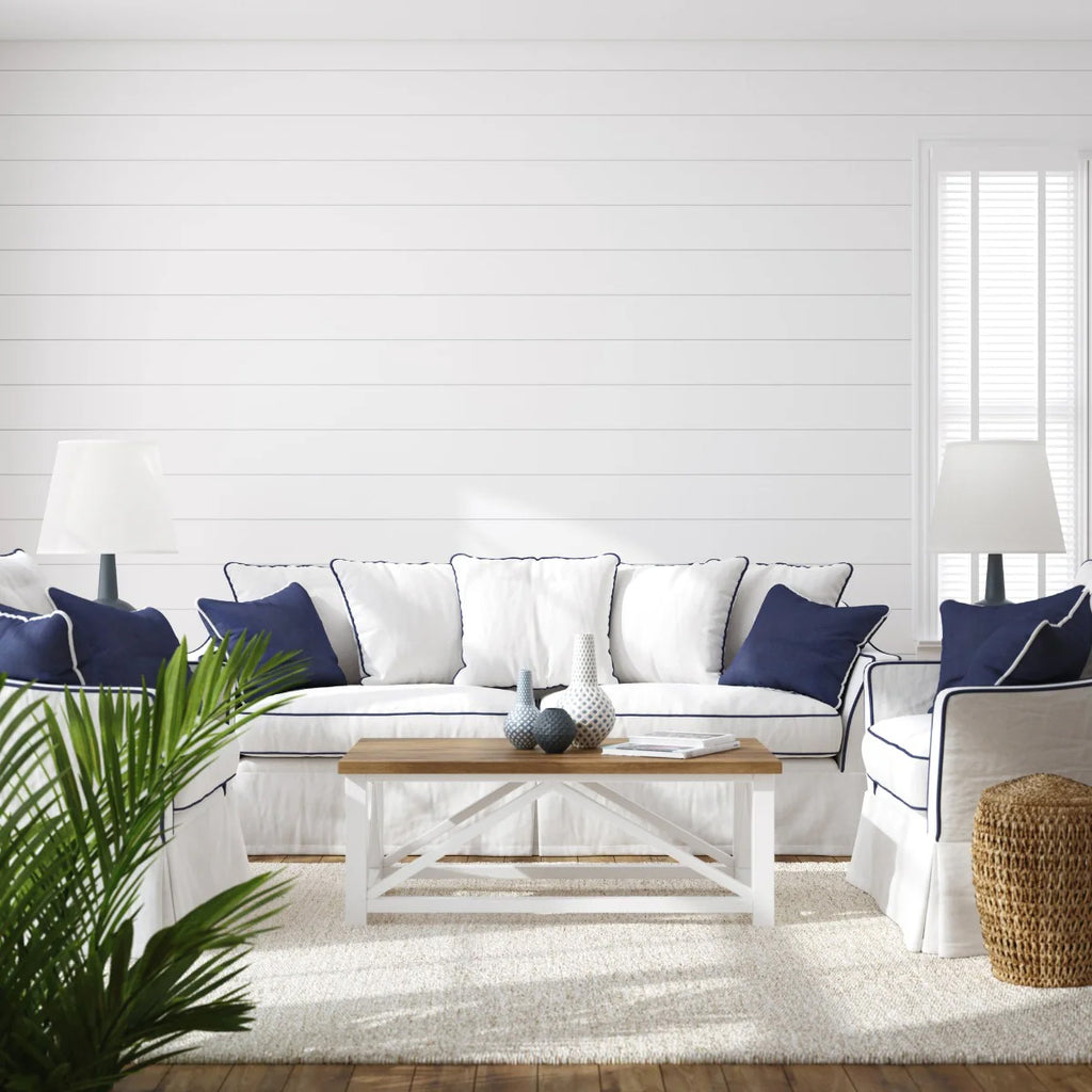 A coastal-style living room bathed in natural light. The white sofa with navy blue and white pillows is the centerpiece. Accents include a white coffee table, table lamps, a woven basket, and potted plants. The room’s charm is enhanced by the Shiplap, Horizontal, Wallpaper in White.
