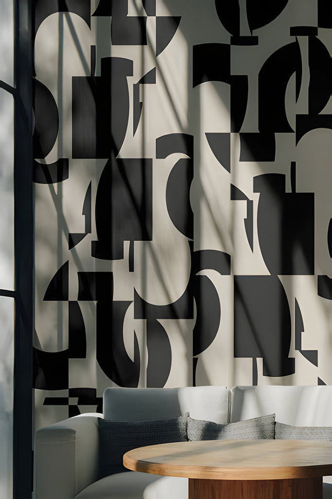 A serene room with sunlight casting shadows on a wall adorned with Slow Assembly Geometric Pattern Wallpaper in Black. The abstract geometric patterns create an intriguing focal point. A cozy corner features a plush sofa and a wooden round table, evoking a comfortable ambiance.