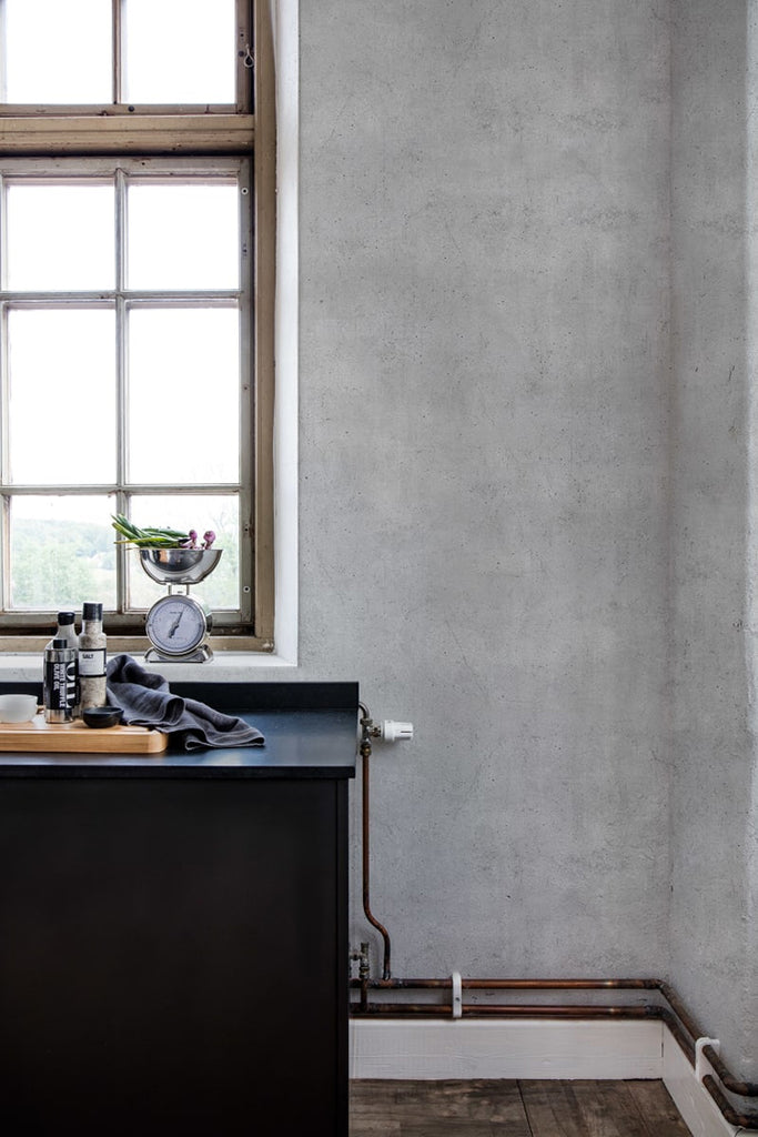 A well-lit room with a large window and wooden floor. The walls are covered with a Solid Concrete, Grey Faux Texture Wallpaper. A dark cabinet with various items is visible.