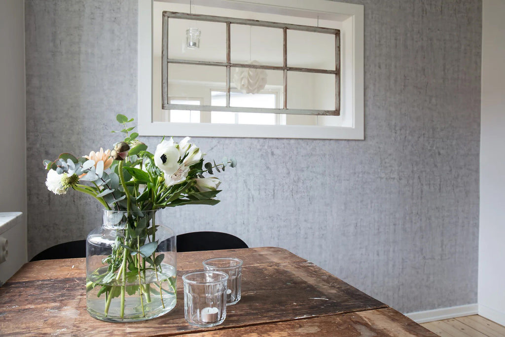 A rustic wooden table with clear glasses and a floral arrangement sits in a cozy room with Solid Concrete, Grey Faux Texture Wallpaper