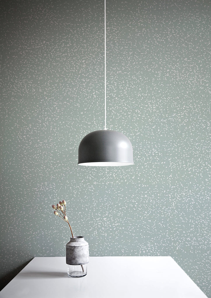 Speckle, Pattern Wallpaper in Sage featured on a wall with white table, a vase with a plant, an a silver pendant light on top