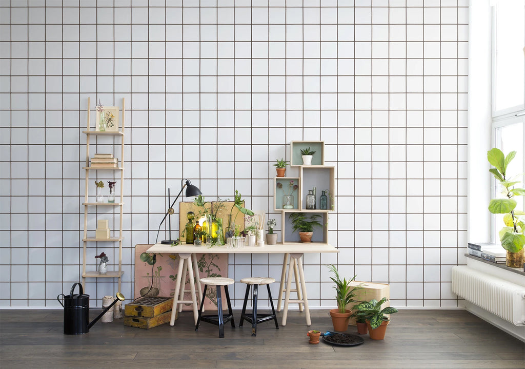 Squares and Tiles illustrations, Wallpaper in White featured on a wall of a room with wooden table and a pair stool surrounded by plants.