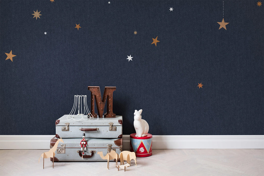 Stargazing, Pattern Wallpaper in dark blue as seen on a wall of a room with stacked briefcase and surrounded by toys