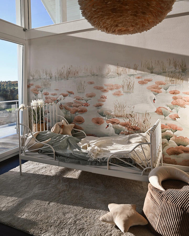 A cozy, sunlit room with a large window. The wall is adorned with Swans and Lilies, Nature Mural Wallpaper in Terracotta. A white metal-framed daybed, a fluffy pendant light fixture, and a striped fabric basket add to the inviting space.