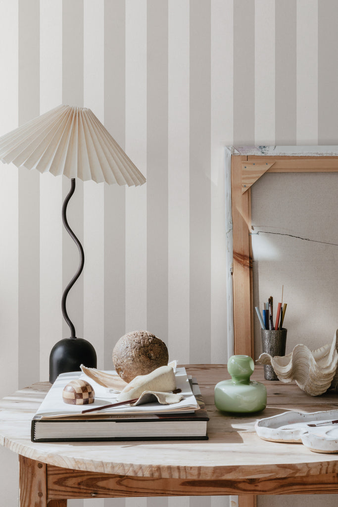 Teira Stripes, Wallpaper in Grey Featured on a wall of a room with wooden round table with books, lamp, and several things on top of it