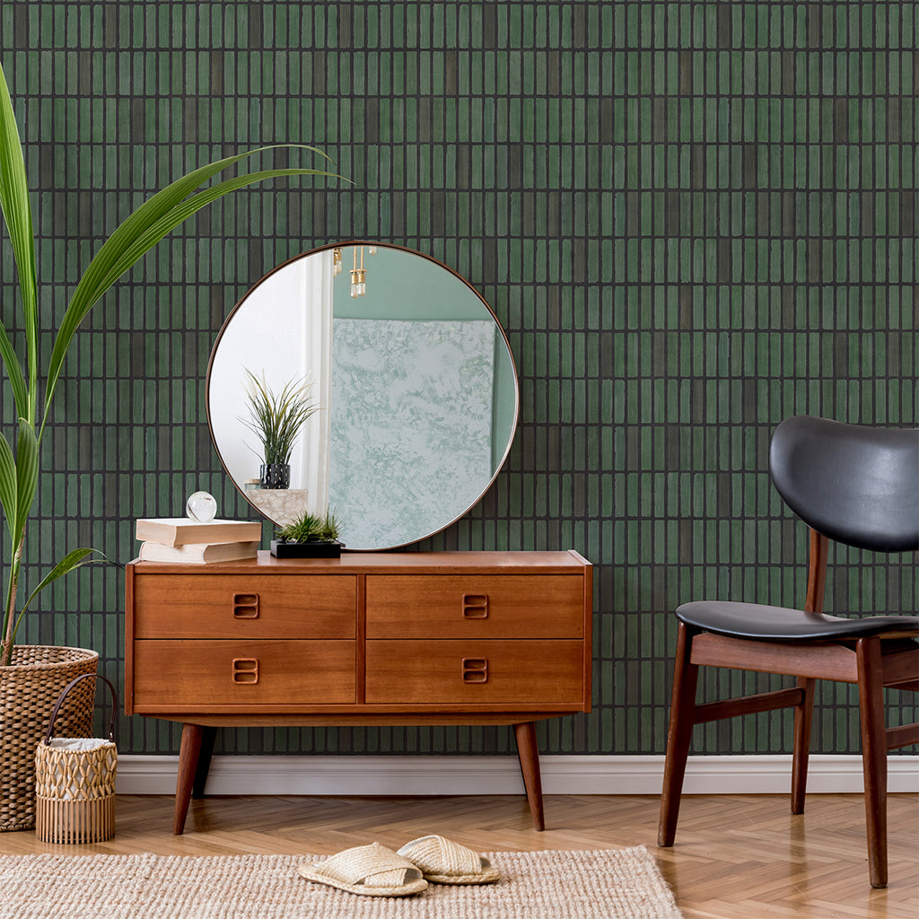 A tastefully decorated mid-century modern room, featuring a wooden sideboard with six drawers, a round mirror above it, and a dark chair to the right. A woven basket with a large green plant adds a touch of nature to the left. The room is enhanced by the Terra Tessel patterned wallpaper in Green, adding a unique texture and visual interest to the space.