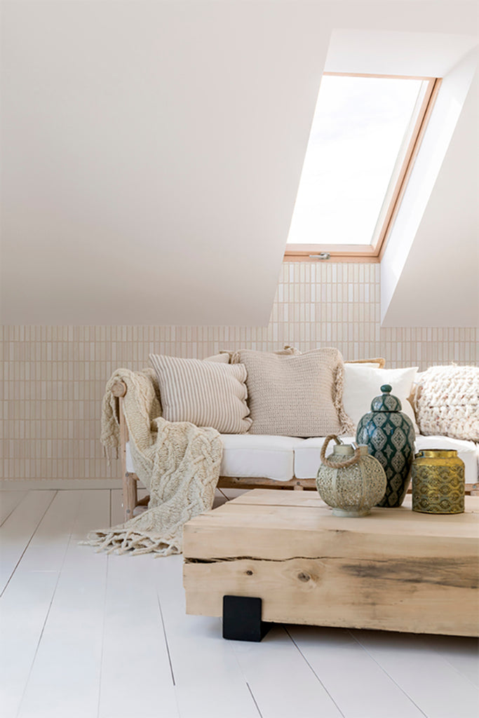A well-lit attic room with a minimalist design. A wooden pallet couch with cream cushions offers a cozy seating area. The room’s serene ambiance is enhanced by the skylight and Terra Tessel, Pattern Wallpaper in Beige.