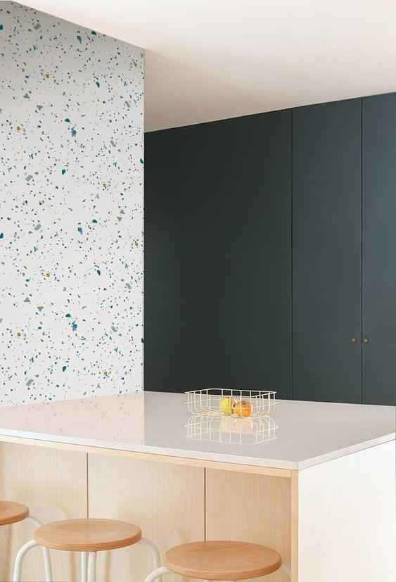 Terrazzo Kie, Wallpaper in blue is showcased on a wall of a kitchen nook. The area features a white finished countertop adorned with fruits, and a wooden high stool.