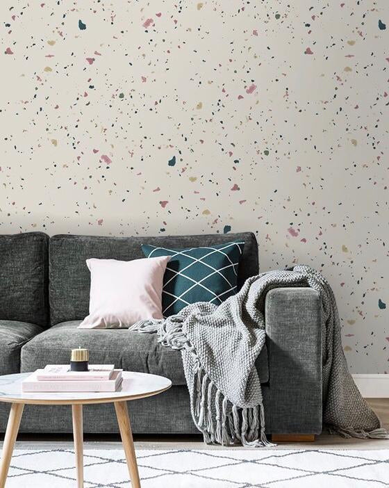 The living area is highlighted by a wall featuring Terrazzo Kie Wallpaper in Sand, a grey sofa with matching sheets and multiple pillows, and a marble-textured table in front, adorned with stacked books.