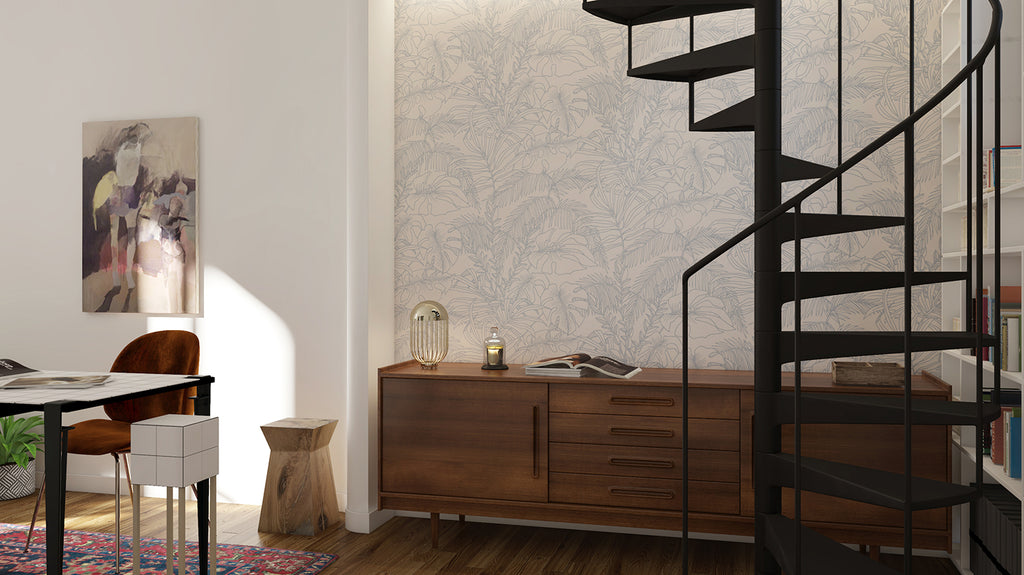 Modern room with Terre, Tropical Pattern Wallpaper in Sand. Features a wooden desk, with books on top, a black spiral staircase, and an abstract painting hanging on the wall.