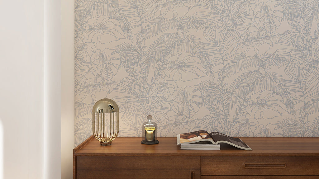 Serene room with Terre, Tropical Pattern Wallpaper in Sand, featuring intricate leaf designs. A wooden cabinet with a golden lamp and a small lantern with a lit candle complements the decor.
