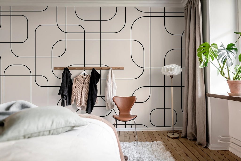 Thea Graphic lines, Pattern Wallpaper Beige featured on a wall of a bedroom with a part of the bed seen and several clothes hanging on the wall. 